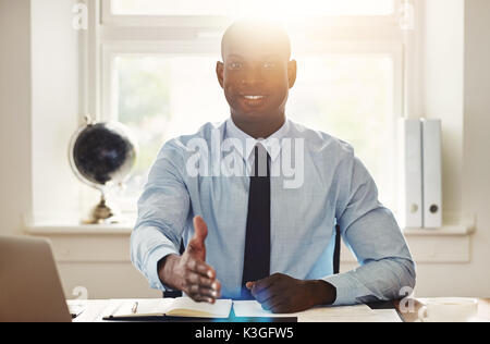 Lächelnden jungen afrikanischen Geschäftsmann, die heraus für einen Händedruck, während an seinem Schreibtisch in einem Büro sitzen Stockfoto