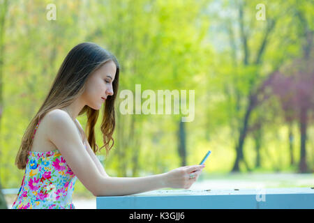 Porträt der jungen schönen Frau mit langem Haar im Sommer Park, Mädchen mit Blume kurzes Kleid mit einem -Smartphone beim Sitzen auf einer Bank Stockfoto