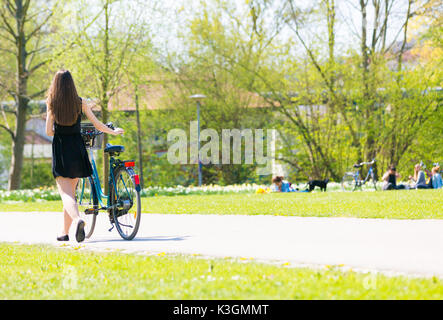 Rückansicht der Mädchen auf dem Fahrrad tragen am Schwarzen kurzen Kleid. Junge Frau Reiten entlang der Straße am grünen Frühling im Park. Sportlich junges Mädchen reiten ein Bic Stockfoto