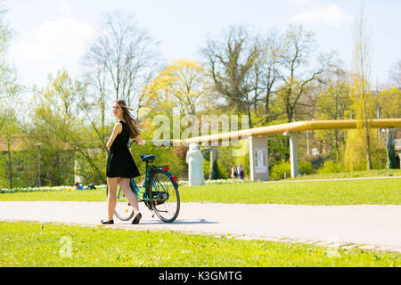 Rückansicht der Mädchen auf dem Fahrrad tragen am Schwarzen kurzen Kleid. Junge Frau Reiten entlang der Straße am grünen Frühling im Park. Sportlich junges Mädchen reiten ein Bic Stockfoto