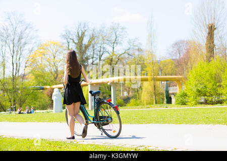 Rückansicht der Mädchen auf dem Fahrrad tragen am Schwarzen kurzen Kleid. Junge Frau Reiten entlang der Straße am grünen Frühling im Park. Sportlich junges Mädchen reiten ein Bic Stockfoto