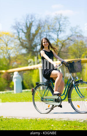 Hochformat von Mdchen auf dem Fahrrad tragen am Schwarzen kurzen Kleid.  Junge Frau Reiten entlang der Strae am grnen Frhling im Park. Sportlich  junges Mdchen ri Stockfotografie - Alamy