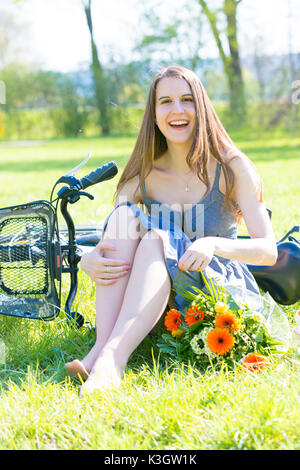 Junge Frau sitzt auf dem Rasen in einem Park in der Nähe ein Fahrrad und ein Blumenstrauß Stockfoto