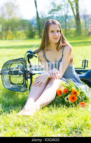 Junge Frau sitzt auf dem Rasen in einem Park in der Nähe ein Fahrrad und ein Blumenstrauß Stockfoto