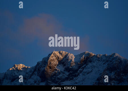 Die westliche Karwendelspitze mit Gipfelkreuz in den letzten winterlichen Abend licht, alpenglühen mit hellen Wolken. Stockfoto