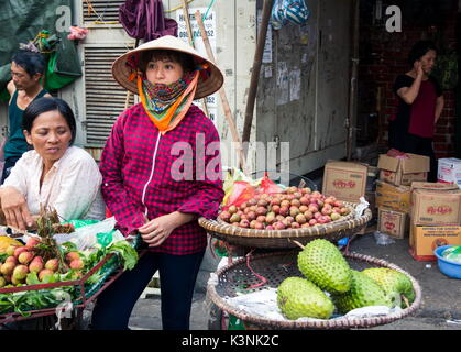 HANOI, VIETNAM - 22. MAI 2017: Frau Verkauf von tropischen Früchten vom Fahrrad Stände auf einem langen Hanoi Street Obstmarkt tragen Vietnamesische konischen Hüten. Stockfoto