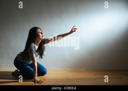 Depression lady kniend auf Holzboden Boden und Hand in den leeren Bereich auf der suche Hilfe auf der weißen Wand Hintergrund mit zerschlagenen missbraucht Licht Stockfoto