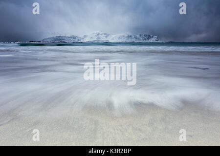 Wellen von der eisigen Meer am Strand im Hintergrund die schneebedeckten Gipfel Ramberg Lofoten Inseln Norwegen Europa Stockfoto