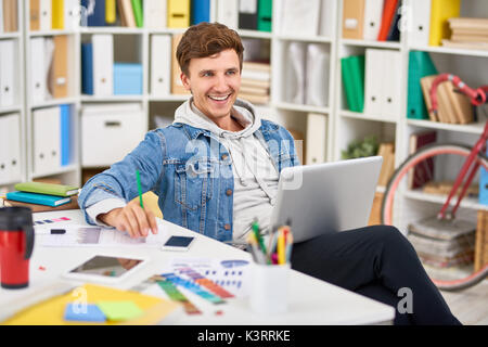 Portrait von lächelnden jungen Mann in Freizeitkleidung mit Laptop, während im modernen Büro gekleidet Stockfoto