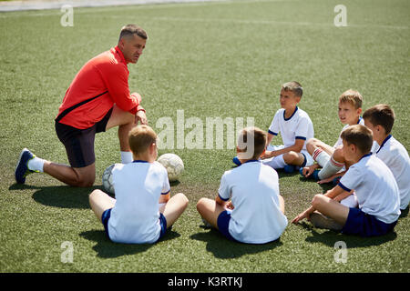 Porträt der jungen Mannschaft sitzen vor dem Trainer auf Fußballfeld zuhören Stockfoto