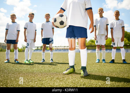 Niedrige Abschnitt Portrait von Fußball-Kapitän steht in der Mitte des Feldes holding Ball mit dem Rest der Mannschaft im Hintergrund Stockfoto