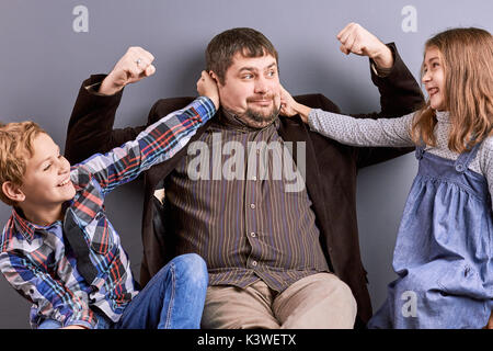 Freundliche Vater spielen mit Kindern. Lustige Vater Darstellung der Muskeln an die Kinder. Konzept der glücklichen Kindheit. Lustige Leute auf grauem Hintergrund. Stockfoto