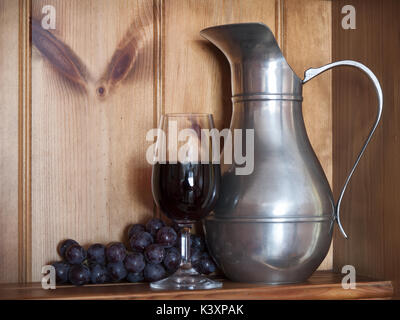 Still Life: Französisch Zinn Krug, Glas Wein, Rotwein, Haufen von schwarzen Trauben auf Eiche Kommode Stockfoto