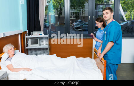 Junge männliche und weibliche Arzt Krankenschwester besuchen und notieren Sie sich die Beschwerden von Patienten, die im Krankenhaus liegt in der Krankenstation. Gesundheitswesen Stockfoto