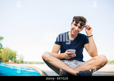 Ein Foto der jungen, gutaussehenden Mann sitzen auf dem Pier und über Handy. Er ist glücklich lächelnd. Stockfoto