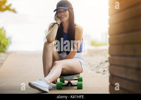 Ein Foto der jungen, schönen Frau sitzt auf ihrem Longboard und glücklich lächelnd. Stockfoto