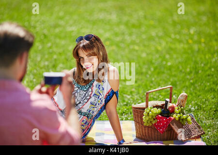 Ein Foto des jungen Paares bei einem Picknick im Park. Der Mann nimmt seine Freundin ein Foto. Stockfoto