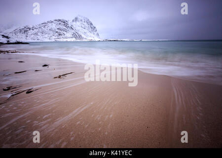 Wellen von der eisigen Meer am Strand im Hintergrund die schneebedeckten Gipfel Pollen Vareid Flakstad Lofoten Inseln Norwegen Europa Stockfoto
