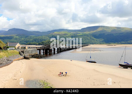 Tywyn, Wales, UK. August 05, 2017. Der hölzerne Eisenbahnbrücke über den Fluss Mawddach mit einem Zug und Urlauber genießen die Sands in der harbo Stockfoto