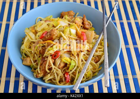 Bami Goreng, gebratene Nudeln mit cabbidge, Sojasauce, Chili, Schweinefleisch in einem blauen Keramik Schüssel auf eine gestreifte Holz Teppich. Stockfoto