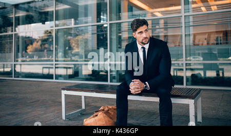 Geschäftsmann sitzt auf der Bank mit Handtasche und Wegsehen. Geschäftsreisende warten außerhalb des Flughafens. Stockfoto