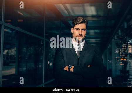 Portrait von stattlicher Junge Unternehmer Stehen mit verschränkten Armen und Kamera. Kaukasische männliche Führungskräfte stehen in einem modernen Gebäude. Stockfoto