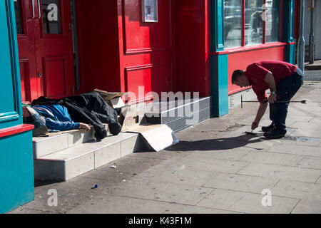 Rough Sleepers in Brighton City Centre. Ein Mann prüft die Wasserversorgung zu einem Geschäft, während jemand in einem Türrahmen schläft. Credit: Terry Applin Stockfoto