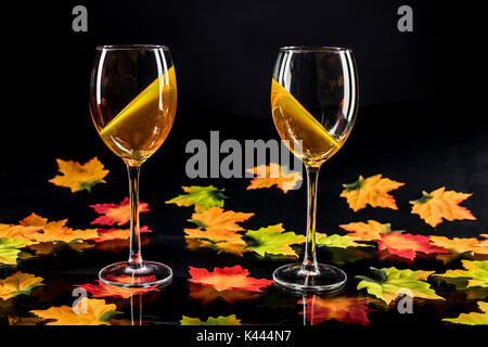 Zwei Weingläser mit Weißwein auf reflektierendem schwarzem Grund mit bunten Weinblättern Stockfoto