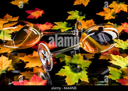 Zwei Weingläser mit Weißwein auf reflektierendem schwarzem Grund mit bunten Weinblättern Stockfoto