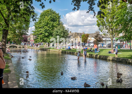 Ein Blick über den Fluss Windrush Besucher genießen den Sommer Sonne auf dem Grün in der Mitte der cotswold Dorf Bourton auf dem Wasser Stockfoto