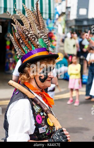 Reifen erwachsenen Frau Volkstänzer von Dead Horse Morris, mit hölzernen Stange auf der Schulter. Von der Seite. Schwarz gemusterten Gesicht Farbe, Motorhaube mit Federn. Stockfoto