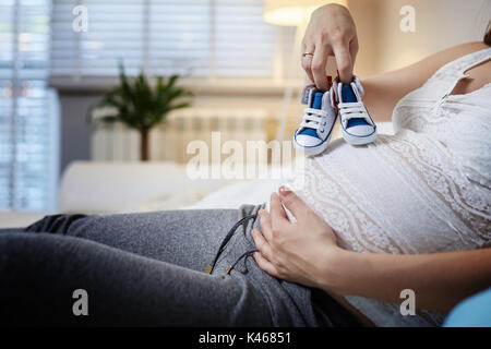 Nicht erkennbare schwangere Frau sitzt auf der Couch und Holding Baby Schuhe. Stockfoto