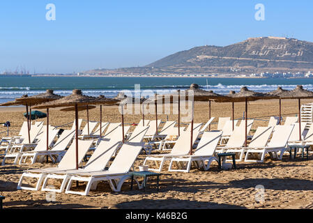 Sonnenschirme und Liegestühle am Strand in Agadir. Marokko. Die Hügel trägt die Inschrift in Arabisch: Gott, Vaterland, König Stockfoto