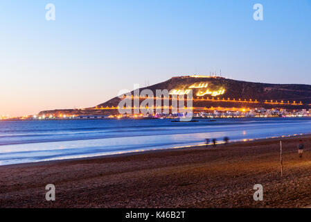 Blick auf langen, breiten Strand in Agadir City bei Nacht, Marokko. Der Berg trägt die Inschrift in Arabisch: Gott, Vaterland, König Stockfoto