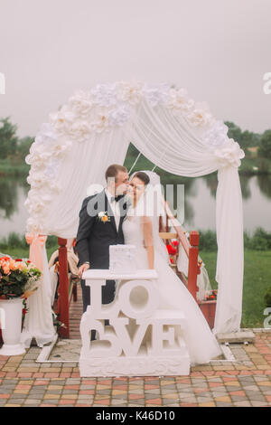 Der Bräutigam ist Küssen die lächelnden Braut in die Wange in der Nähe der Hochzeit Schild und Arch. Stockfoto