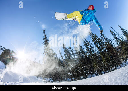 Snowboarder, Freerider das Springen von einem Schnee Rampe in der Sonne auf dem Hintergrund der Wald und die Berge. Stockfoto