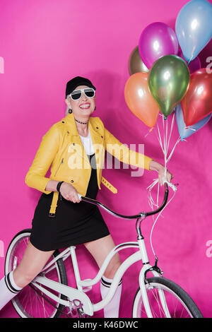 Modische senior Frau tragen gelbe Leder Jacke mit Fahrrad und bunten Ballons isoliert auf Rosa Stockfoto