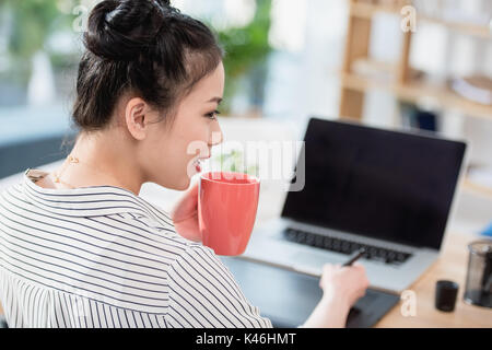 Junge asiatische Designer Kaffee trinken während der Arbeit mit Grafiktablett und Laptop Stockfoto