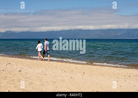 Ein liebevolles Paar von einem Mann und einer Frau Spaziergänge entlang der Küste Sand des Baikalsees im Sommer während einer Tide auf einem Berg im Hintergrund an einem klaren Sonne Stockfoto