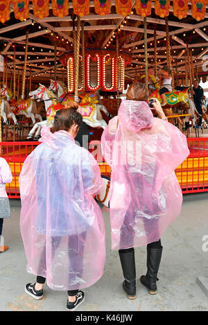 Zwei Frauen mit dem Rücken zur Kamera, die pinkfarbene Umhänge tragen und vor dem traditionellen Karussell stehen, drehen sich um, wenn der Regen anfängt Stockfoto