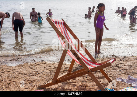 Einen leeren Liegestuhl am Strand mit Leuten spielen im Meer im Hintergrund. Southend-on-Sea, Essex, Großbritannien Stockfoto