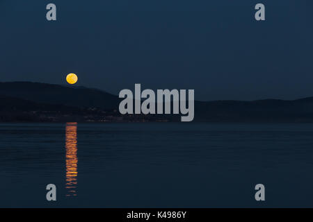 Vollmond über einige Hügel auf einem See, perfekt auf dem Wasser widerspiegeln Stockfoto