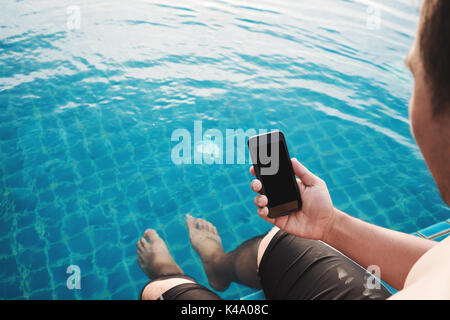 Close-up ein Mann mit smart phone mit leeren Kopie Platz auf dem Bildschirm, mit Beinen Tauchen im Pool im Sommer für Urlaub. Beschneidungspfade auf dem Bildschirm Stockfoto