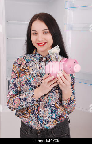 Junge schöne Frau mit Sparschwein (Spardose) auf dem Kühlschrank Hintergrund Stockfoto