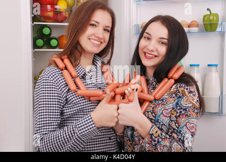 Zwei junge Mädchen hält Würstchen auf dem Kühlschrank Hintergrund. Zwei schöne junge Mädchen in der Nähe der Kühlschrank. Stockfoto