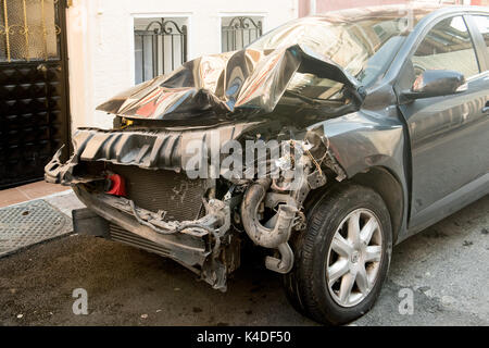 Ein schwer beschädigtes schwarzes Auto mit einer zerknitterten Front und freiliegenden Motorteilen, das nach einem Unfall auf einer Wohnstraße geparkt wurde. Stockfoto