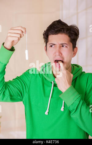 Stattlicher Mann mit ein angewidertes Gesicht eine Menstruation Baumwolle Tampon, tut ein Zeichen mit dem Finger in den Mund und trug eine grüne Hoodie in einem verschwommenen Hintergrund Stockfoto