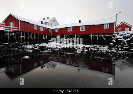 Starker Schneefall über den roten Rorbuer - einmal traditionelle saisonale Fischerhütten - Jetzt für touristische Nutzung auf dem eisigen Wasser spiegelt - Hafen von Hamnoy Dorf in der Nähe von Lauf Stockfoto