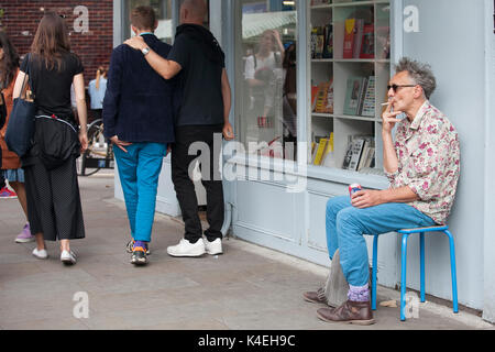 LONDON, ENGLAND - 20. August 2017 Ein alter grauhaariger Mann sitzt auf einem Stuhl in der Nähe der Wand und raucht am Broadway Road Market Stockfoto