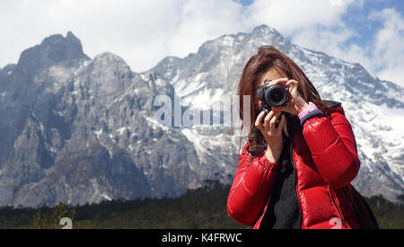 Junge Mädchen Fotograf holding Nikon DSLR, sie Kraft auf andere, mit Jade Dragon Snow Mountain Hintergrund in Lijiang, im Winter, in Yunnan China Breite Stockfoto